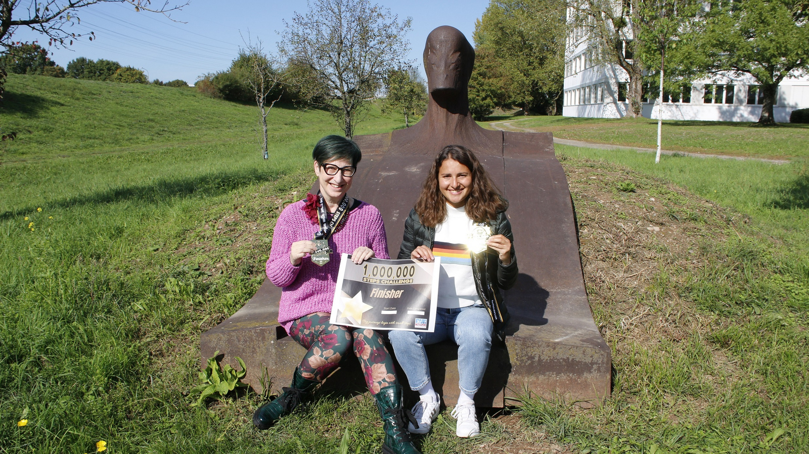 Cornelia Herde und Stella Schneider (von links) sitzen auf der liegenden Eisenskulptur an der Hochschule und halten ihre Finisher-Medaillen und eine Urkunde in die Kamera