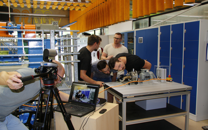 Studierende beugen sich neugierig über eine Maschine und werden dabei von einem KAmermann gefilmt