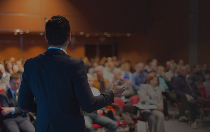 Ein Referent steht mit dem Rücken zur Kamera vor einem sitzenden, nur unscharf zu erkennenden Publikum 