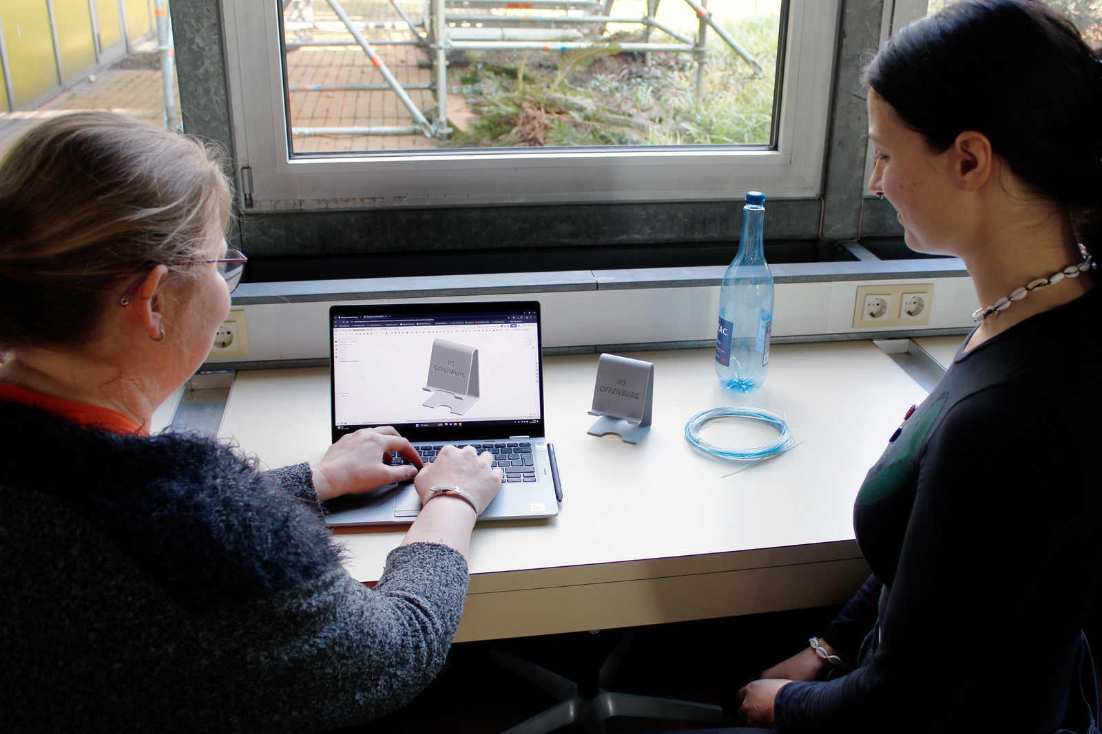 zwei junge Frauen sitzen an einem Laptop auf dem eine Konstruktionsskizze eines Smartphone-Halters zu sehen ist. Neben ihnen auf dem Tisch eine leere Plastikflasche, Filament und ein fertiger Smartphone-Halter aus dem 3D-Drucker
