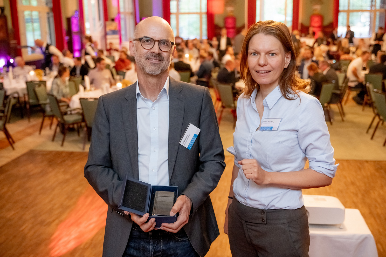 Tobias Hagen und Nicole Ondrusch stehen nebeneinander in einem Saal, im Hintergrund sind verschwommen die sitzenden Konferenzteilnehmer zu erkennen, Tobias HAgen hält die Schatulle mit der medaille in den Händen