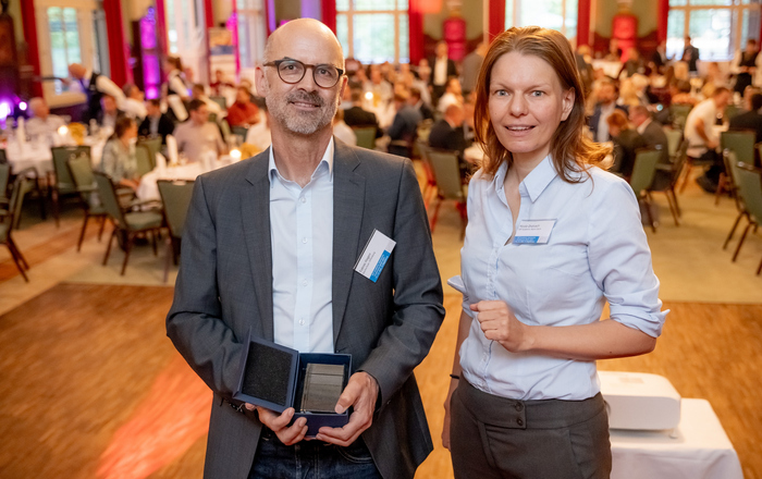 Tobias Hagen und Nicole Ondrusch stehen nebeneinander in einem Saal, im Hintergrund sind verschwommen die sitzenden Konferenzteilnehmer zu erkennen, Tobias HAgen hält die Schatulle mit der medaille in den Händen