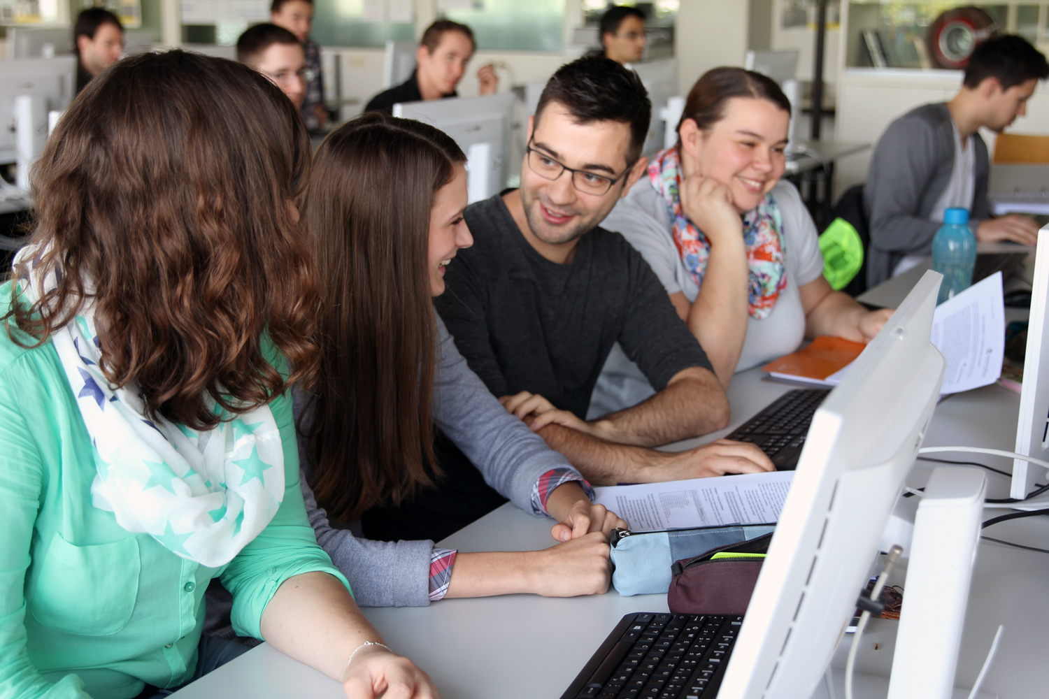 Lachende weibliche und männliche Studierende sitzen vor PC-Bildschirmen und unterhalten sich.