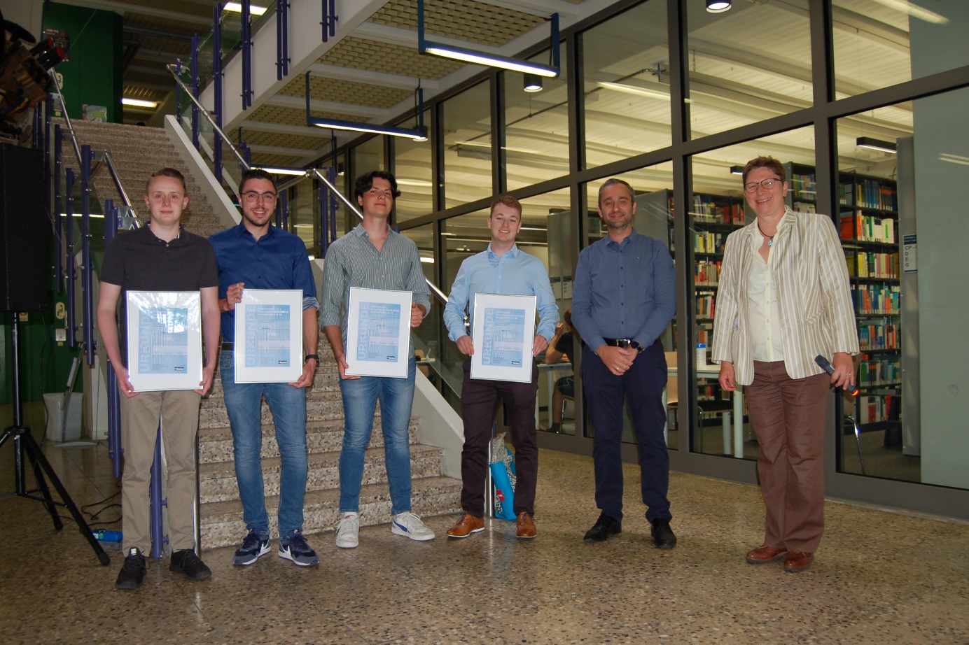 Die Preisträger, Firmenvertreter Alexander Becker und Prof.in Elke Mackensen (von links) stehen im Foyer von Gebäude B vor einer Treppe
