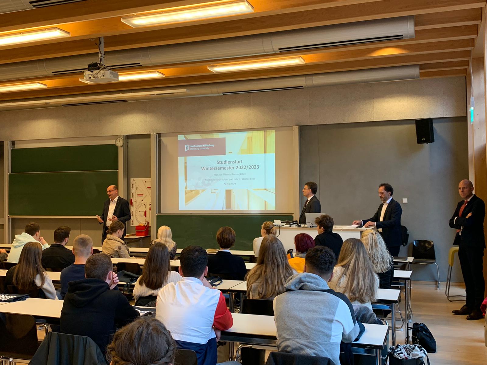 Rektor Stephan Trahasch, Bürgermeister Thorsten Erny und die professoren Thomas Baumgärtler und Andreas Klasen stehen in der Aula vor den sitzenden Erstsemestern hinter ihnen ist auf einer Leinwand die Startseite einer Powerpoint-Präsentatio zu sehen