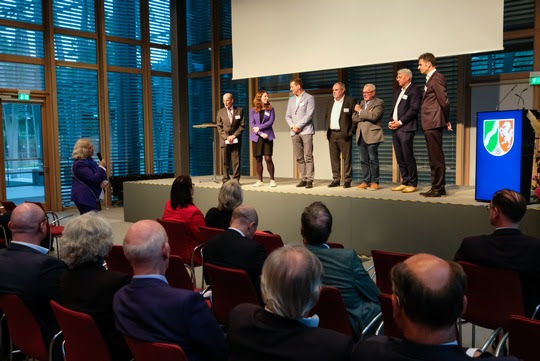 Eine Gruppe von Leuten steht auf einer Bühne vor sitzenden Zuhörenden