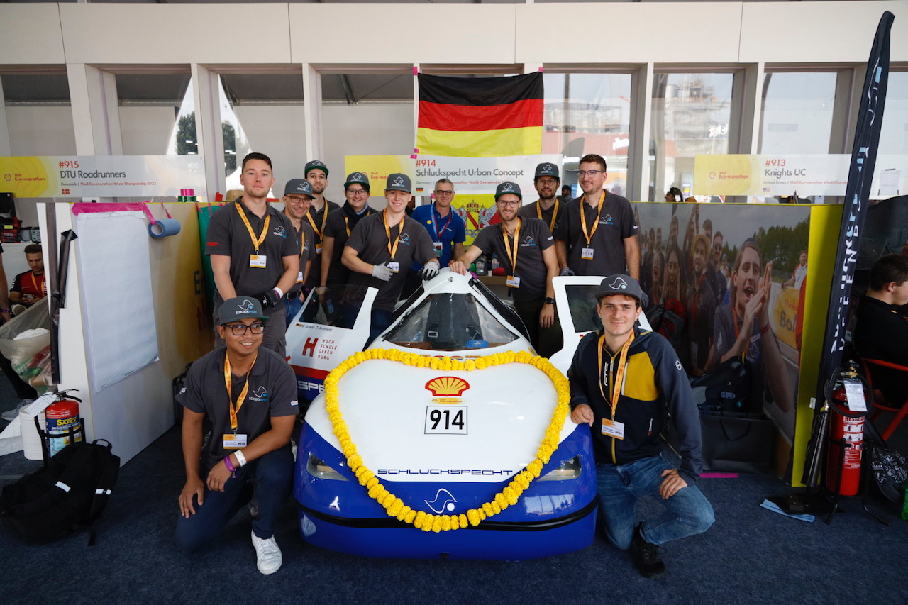 Team steht um das mit einem gelben Blumenkranz geschmückte Auto herum