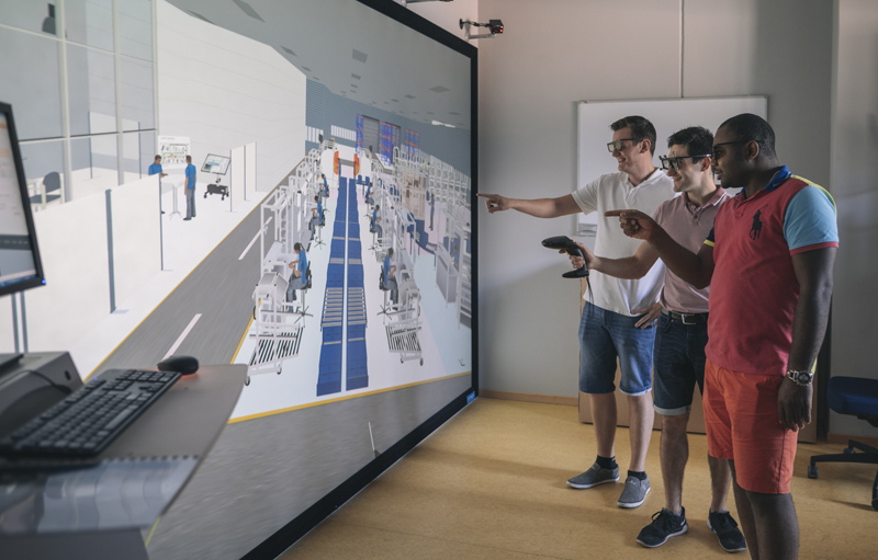 drei Personen stehen vor einem großen Bildschirm auf dem eine virtuelle Fabrik zu sehen ist